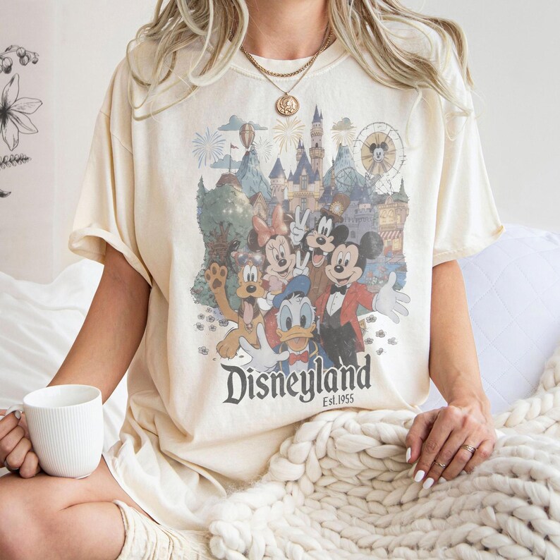 May include: Cream-colored t-shirt featuring a Disneyland graphic with Mickey Mouse, Minnie Mouse, Donald Duck, Goofy, and Pluto. The graphic includes a castle, fireworks, and a Ferris wheel. The text "Disneyland Est. 1955" is printed below the characters.
