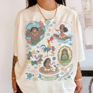 May include: Cream-colored t-shirt featuring colorful illustrations of characters from the animated film "Moana." The design includes images of Maui, Moana, Pua, Hei Hei, and other elements from the movie, with the text "Moana & Friends."