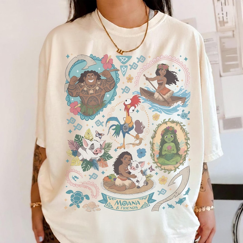May include: Cream-colored t-shirt featuring colorful illustrations of characters from the animated film "Moana." The design includes images of Maui, Moana, Pua, Hei Hei, and other elements from the movie, with the text "Moana & Friends."