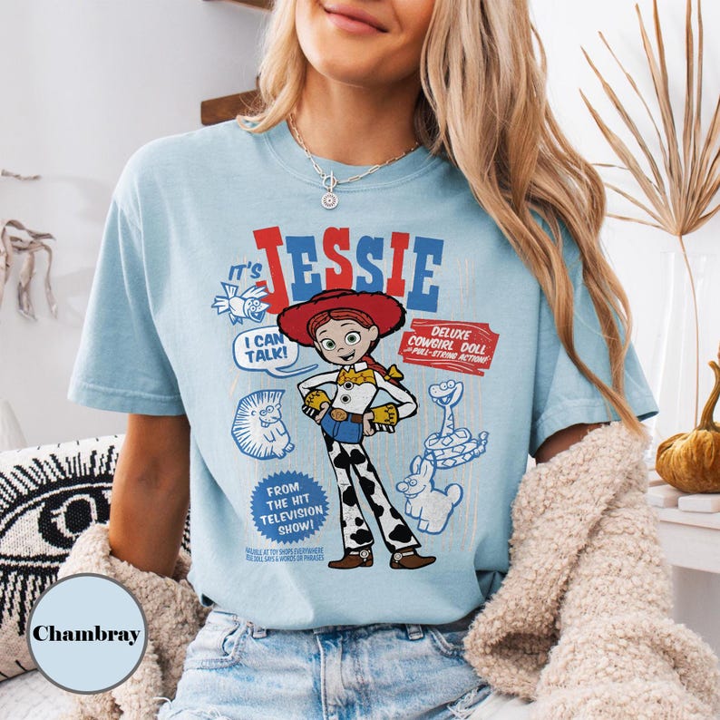 May include: Light blue t-shirt featuring a graphic of Jessie from Toy Story. The design includes the text "It's Jessie" in red and blue, along with cartoon illustrations and the phrases "I Can Talk!" and "Deluxe Cowgirl Doll".