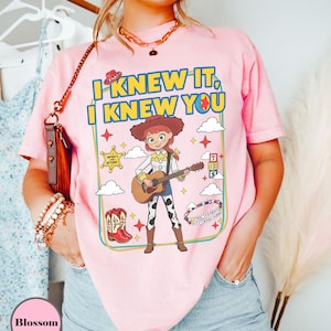 May include: A light pink t-shirt featuring a graphic print of a cowgirl character playing an acoustic guitar. The text reads I KNEW IT, I KNEW YOU. The design includes a sheriff badge, red cowboy boots, alphabet blocks, and clouds within a frame.