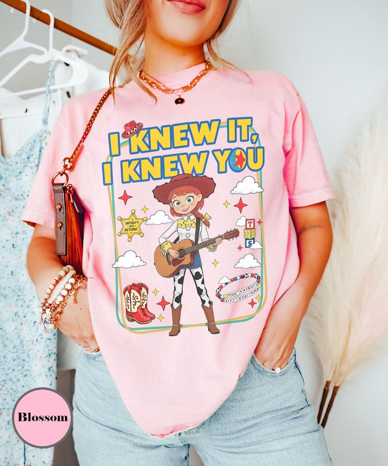 May include: A light pink t-shirt featuring a graphic print of a cowgirl character playing an acoustic guitar. The text reads I KNEW IT, I KNEW YOU. The design includes a sheriff badge, red cowboy boots, alphabet blocks, and clouds within a frame.