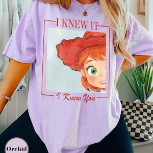 K&ouml;nnte beinhalten: Ein hellviolettes Kurzarm-T-Shirt mit einer rot umrandeten quadratischen Grafik. Im Quadrat befindet sich eine Aquarell-Illustration einer Figur mit roten Haaren und einem roten Cowboyhut. Der Text I KNEW IT und I Knew You ist in Rot gedruckt.