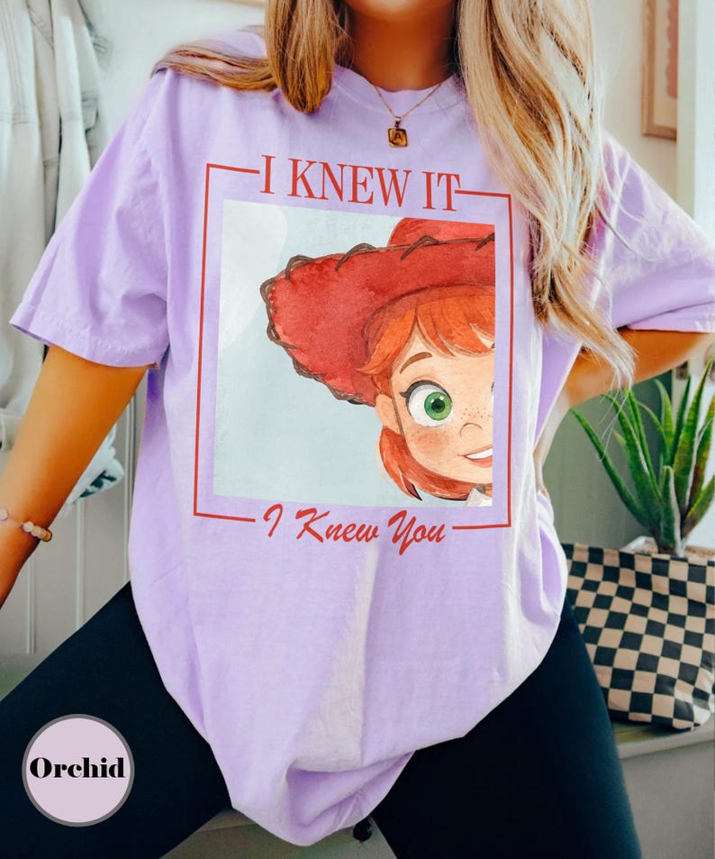 K&ouml;nnte beinhalten: Ein hellviolettes Kurzarm-T-Shirt mit einer rot umrandeten quadratischen Grafik. Im Quadrat befindet sich eine Aquarell-Illustration einer Figur mit roten Haaren und einem roten Cowboyhut. Der Text I KNEW IT und I Knew You ist in Rot gedruckt.