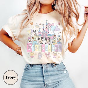 May include: Ivory-colored t-shirt featuring a colorful Disneyland graphic. The design includes a castle, Mickey and Minnie Mouse, and other Disney characters. The word "Disneyland" is displayed in pastel colors.