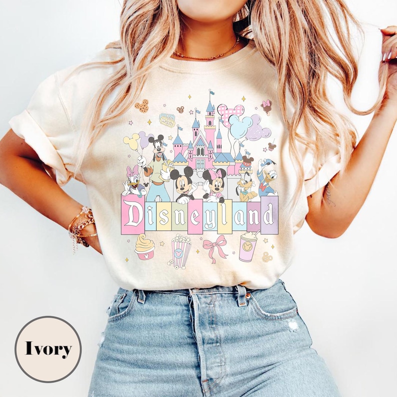 May include: Ivory-colored t-shirt featuring a colorful Disneyland graphic. The design includes a castle, Mickey and Minnie Mouse, and other Disney characters. The word "Disneyland" is displayed in pastel colors.
