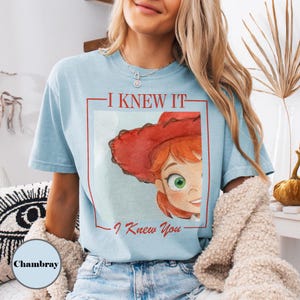 K&ouml;nnte beinhalten: Ein hellblaues T-Shirt mit einem Grafikdruck einer Zeichentrickfigur mit roten Haaren und einem roten Cowboyhut. Der Text "I KNEW IT" steht &uuml;ber der Figur und "I Knew You" darunter, alles eingerahmt in einem roten rechteckigen Rahmen.