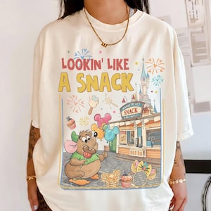 May include: Cream-colored t-shirt with the phrase "Lookin' Like A Snack" in yellow and red. The graphic features a cartoon mouse holding a lollipop, balloons, and a snack stand with a castle backdrop. Colorful fireworks and food illustrations complete the design.