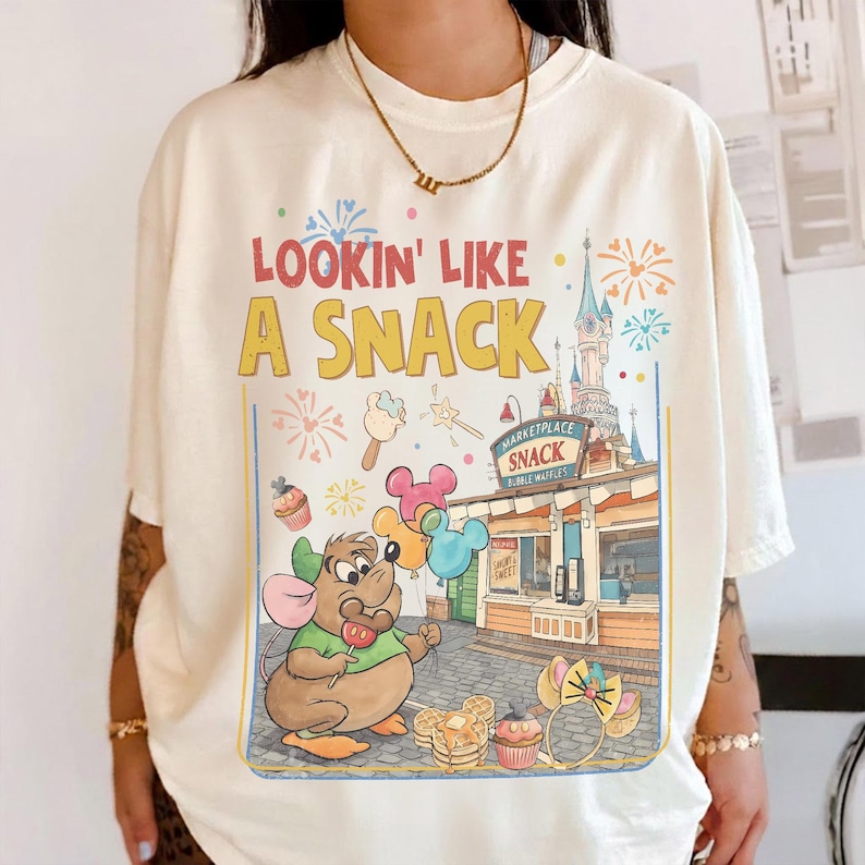 May include: Cream-colored t-shirt with the phrase "Lookin' Like A Snack" in yellow and red. The graphic features a cartoon mouse holding a lollipop, balloons, and a snack stand with a castle backdrop. Colorful fireworks and food illustrations complete the design.