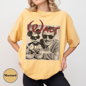 May include: Mustard-colored t-shirt featuring a vintage graphic of Mickey Mouse and Walt Disney wearing sunglasses. The word "Walt" is printed in red above the image. The shirt is a relaxed fit.
