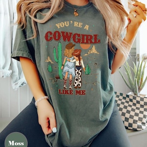 May include: A moss green short-sleeve t-shirt featuring a graphic of two figures in cowboy hats standing in a desert landscape with cacti and mountains. The text reads "YOU'RE A COWGIRL LIKE ME" in red and yellow lettering across the front of the shirt.
