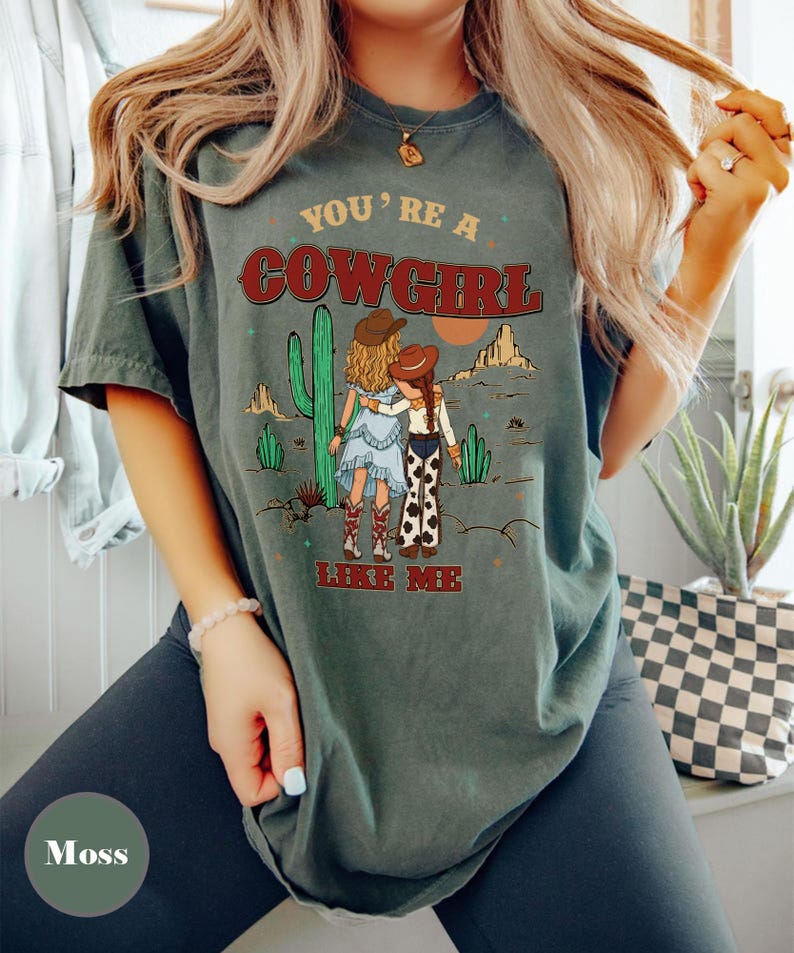 May include: A moss green short-sleeve t-shirt featuring a graphic of two figures in cowboy hats standing in a desert landscape with cacti and mountains. The text reads "YOU'RE A COWGIRL LIKE ME" in red and yellow lettering across the front of the shirt.