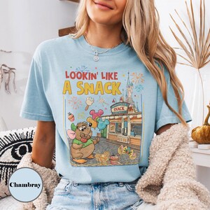 May include: Light blue t-shirt with the phrase "Lookin' Like A Snack" and a cartoon graphic of a mouse holding balloons and a lollipop. The graphic includes a snack shop and fireworks.