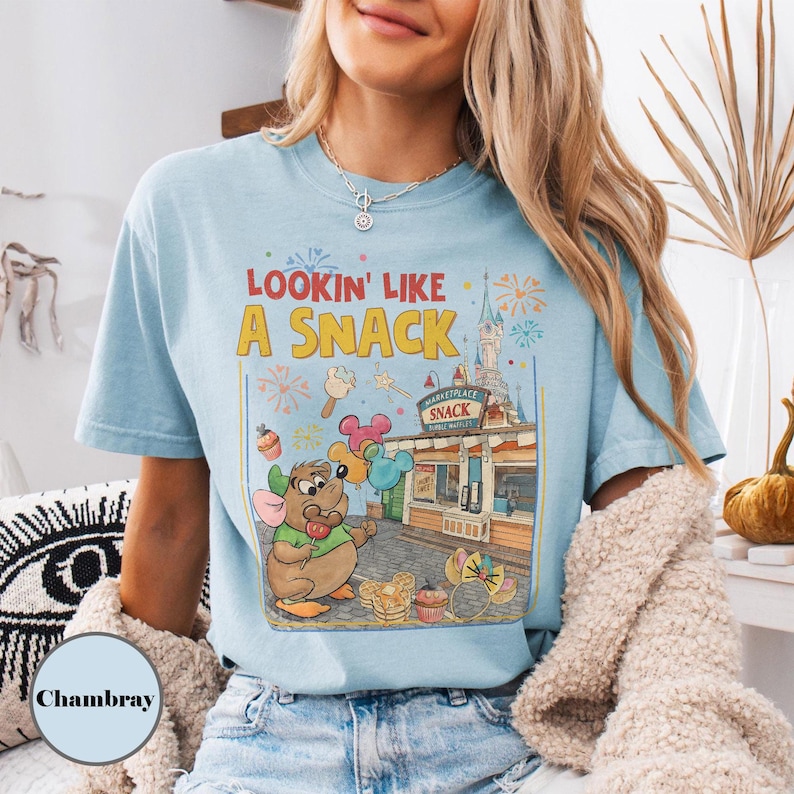 May include: Light blue t-shirt with the phrase "Lookin' Like A Snack" and a cartoon graphic of a mouse holding balloons and a lollipop. The graphic includes a snack shop and fireworks.