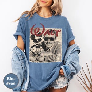 May include: Blue jean-colored t-shirt featuring a vintage graphic of Walt Disney with Mickey Mouse wearing sunglasses. The word "WALT" is printed above the image in a red, stylized font. The shirt has rolled-up sleeves.