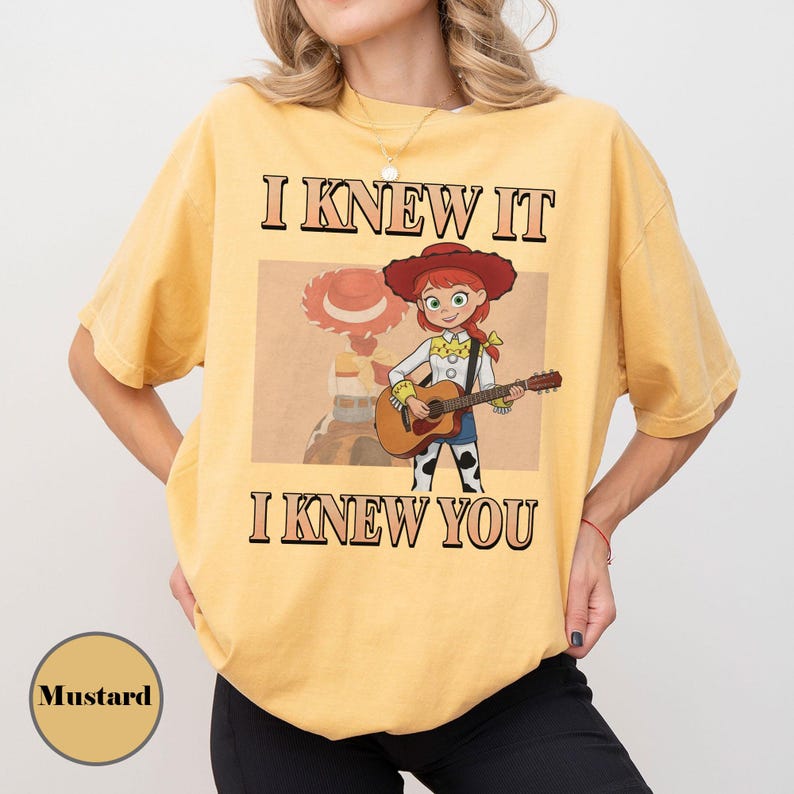 May include: A mustard yellow short-sleeve t-shirt featuring a graphic of a cartoon cowgirl playing an acoustic guitar. The text I KNEW IT and I KNEW YOU is printed in black above and below the illustration. The design includes a tan rectangular background.
