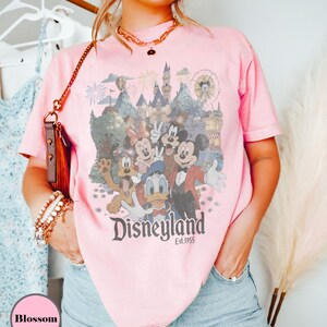 May include: Pink t-shirt featuring a Disneyland graphic with Mickey Mouse, Minnie Mouse, Donald Duck, Goofy, and other characters. The graphic includes a castle, fireworks, and a Ferris wheel. The text "Disneyland Est. 1955" is printed below the characters.