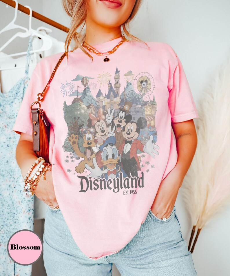May include: Pink t-shirt featuring a Disneyland graphic with Mickey Mouse, Minnie Mouse, Donald Duck, Goofy, and other characters. The graphic includes a castle, fireworks, and a Ferris wheel. The text "Disneyland Est. 1955" is printed below the characters.