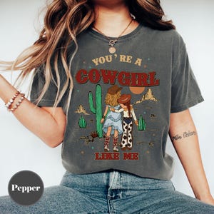 May include: A charcoal gray short-sleeve t-shirt featuring a graphic of two figures in cowboy hats standing in a desert landscape with cacti. The text reads YOU'RE A COWGIRL LIKE ME in red and yellow lettering. The shirt has a crew neckline.