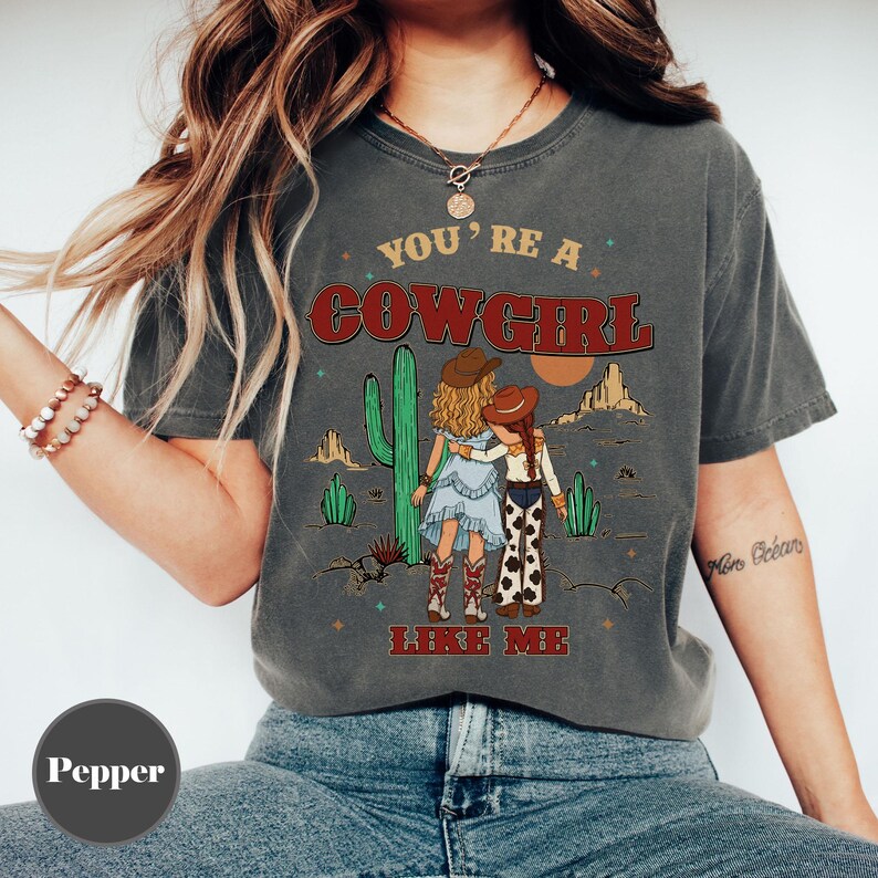 May include: A charcoal gray short-sleeve t-shirt featuring a graphic of two figures in cowboy hats standing in a desert landscape with cacti. The text reads YOU'RE A COWGIRL LIKE ME in red and yellow lettering. The shirt has a crew neckline.