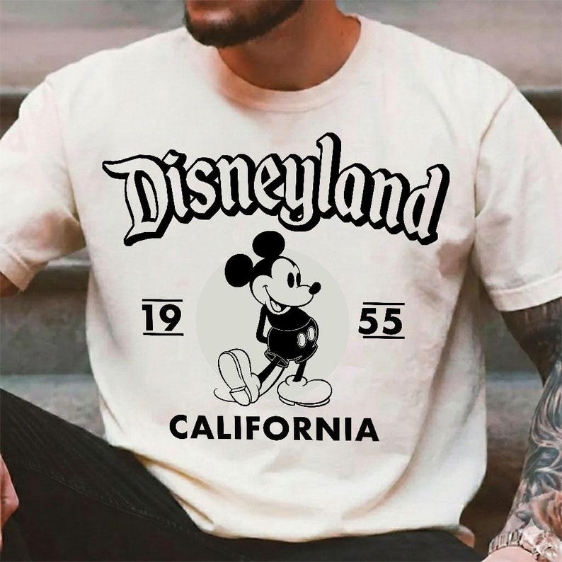 May include: Cream-colored t-shirt featuring the word "Disneyland" in a black, stylized font. Below, a smiling Mickey Mouse graphic is centered within a gray circle, with the numbers "19" and "55" on either side. The word "CALIFORNIA" is printed below.
