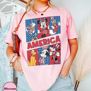 May include: Pink t-shirt featuring a graphic of Disney characters in patriotic attire. The word "AMERICA" is displayed in red and white block letters. The shirt showcases Daisy Duck, Minnie Mouse, Donald Duck, Pluto, Mickey Mouse, and Goofy.