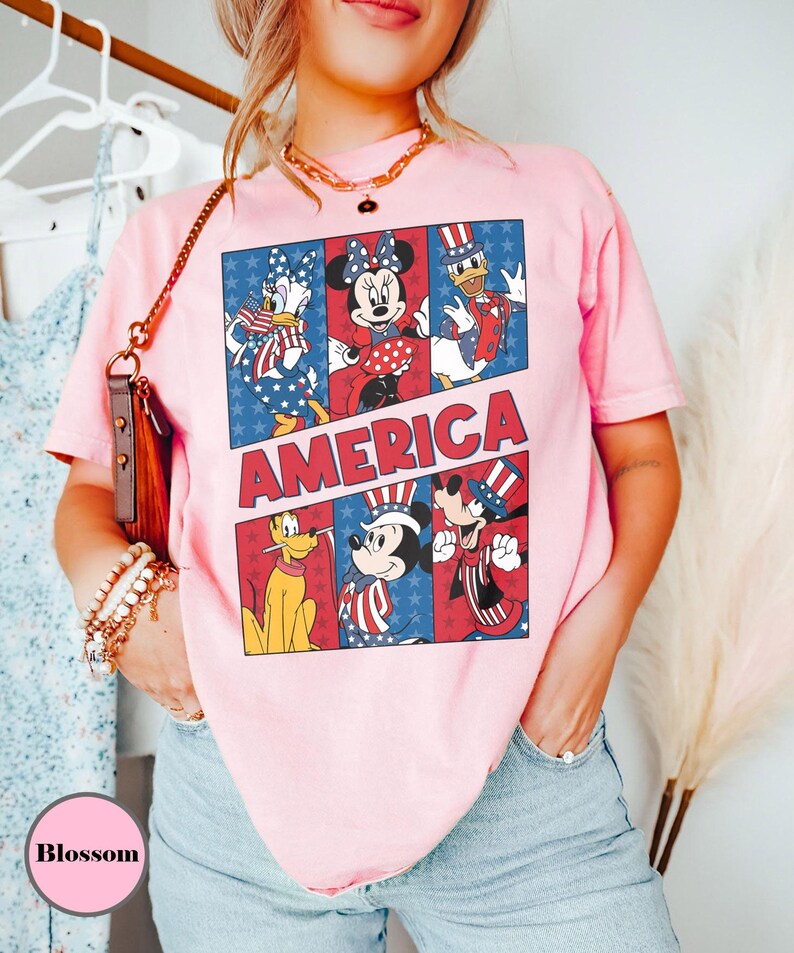 May include: Pink t-shirt featuring a graphic of Disney characters in patriotic attire. The word "AMERICA" is displayed in red and white block letters. The shirt showcases Daisy Duck, Minnie Mouse, Donald Duck, Pluto, Mickey Mouse, and Goofy.