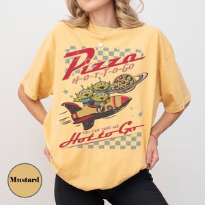 May include: Mustard-coloured t-shirt with a graphic featuring the text "Pizza H-O-T-T-O-GO" and "You Can Take Me Hot to Go." The design includes three green aliens in a rocket ship with a pizza and planet.