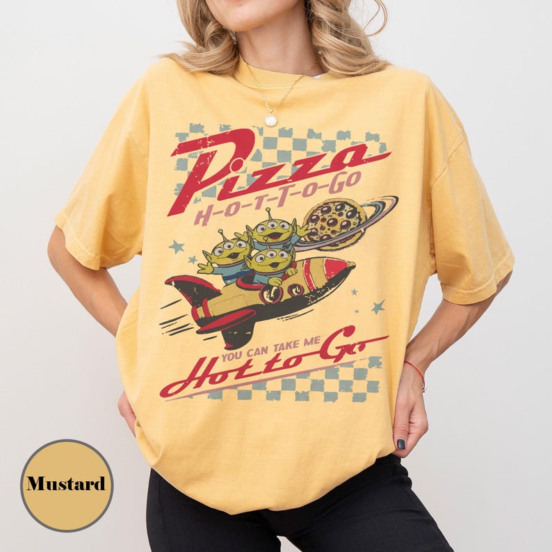 May include: Mustard-coloured t-shirt with a graphic featuring the text "Pizza H-O-T-T-O-GO" and "You Can Take Me Hot to Go." The design includes three green aliens in a rocket ship with a pizza and planet.