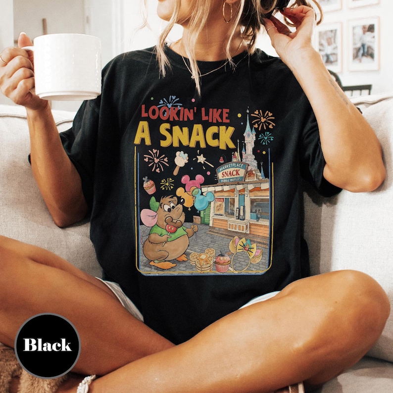 May include: Black t-shirt with a colorful graphic featuring a cartoon mouse, a snack stand, and the text "Lookin' Like A Snack." The design includes fireworks, cupcakes, and balloons. The shirt is a casual wear item.
