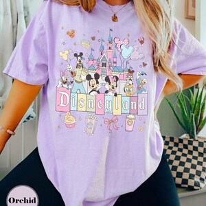 May include: A light purple t-shirt featuring a colorful Disneyland graphic. The design includes the Disneyland castle, Mickey and Minnie Mouse, Donald Duck, and other Disney characters. The word "Disneyland" is displayed in pastel colors.