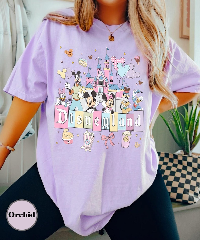 May include: A light purple t-shirt featuring a colorful Disneyland graphic. The design includes the Disneyland castle, Mickey and Minnie Mouse, Donald Duck, and other Disney characters. The word "Disneyland" is displayed in pastel colors.