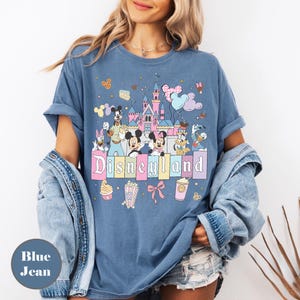 May include: Blue jean t-shirt featuring a Disneyland graphic with cartoon characters and a castle. The design includes Mickey and Minnie Mouse, Donald Duck, and Goofy. The word "Disneyland" is displayed in pastel colors.