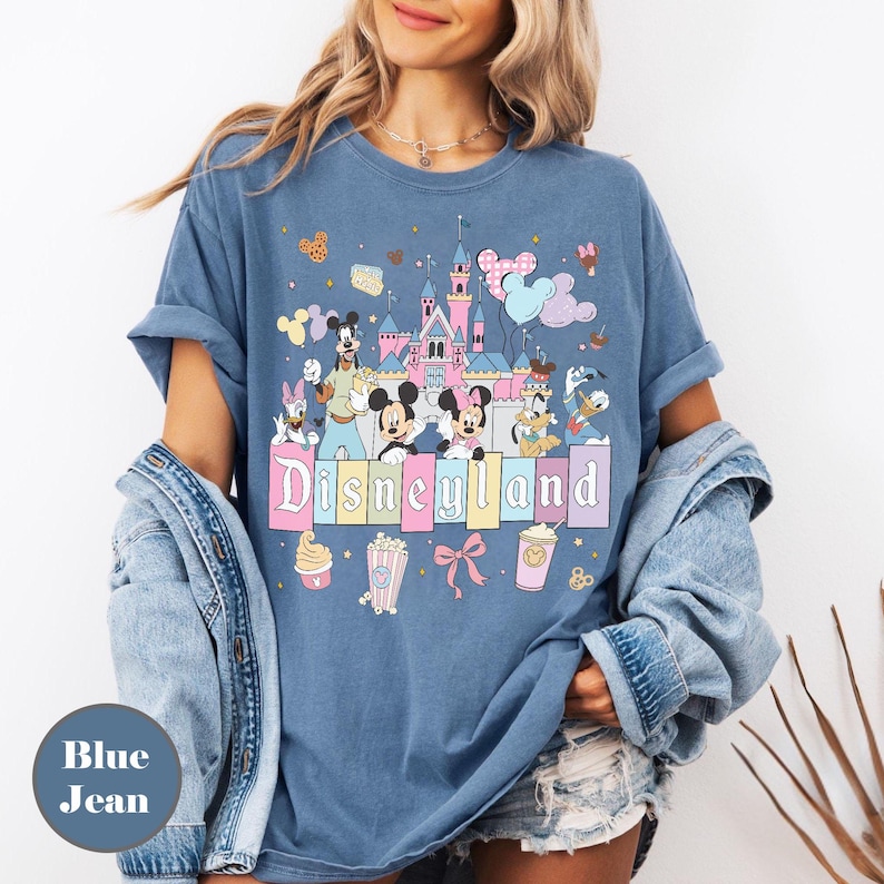 May include: Blue jean t-shirt featuring a Disneyland graphic with cartoon characters and a castle. The design includes Mickey and Minnie Mouse, Donald Duck, and Goofy. The word "Disneyland" is displayed in pastel colors.
