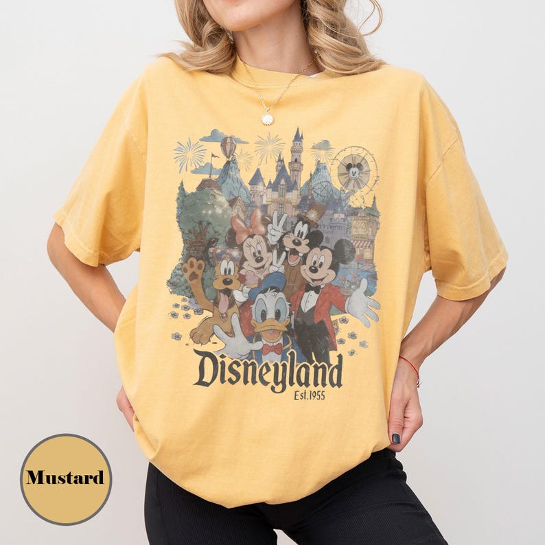 May include: Mustard-colored t-shirt featuring a Disneyland graphic with Mickey Mouse, Minnie Mouse, Donald Duck, and Goofy. The graphic includes a castle, fireworks, and a Ferris wheel. The text "Disneyland Est. 1955" is printed below the characters.