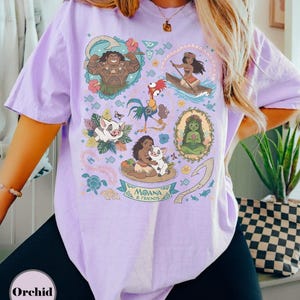 May include: A light purple t-shirt featuring colorful illustrations of characters from the animated film "Moana." The design includes images of Maui, Moana, Pua, Hei Hei, and other elements from the movie. The text "Moana & Friends" is also visible.