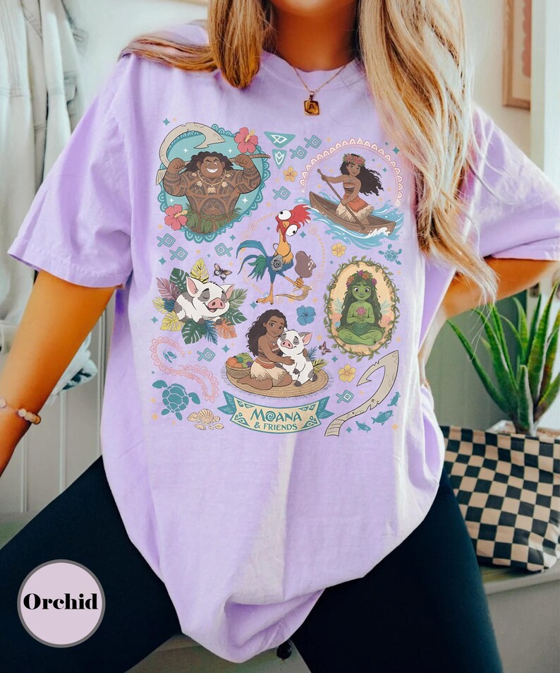May include: A light purple t-shirt featuring colorful illustrations of characters from the animated film "Moana." The design includes images of Maui, Moana, Pua, Hei Hei, and other elements from the movie. The text "Moana & Friends" is also visible.