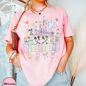 May include: Pink t-shirt featuring a Disneyland graphic with Mickey Mouse, Minnie Mouse, Donald Duck, and other Disney characters. The graphic includes a castle, balloons, and the word "Disneyland" in pastel colors. The shirt is a light pink color.