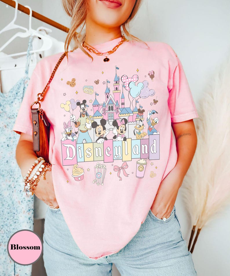 May include: Pink t-shirt featuring a Disneyland graphic with Mickey Mouse, Minnie Mouse, Donald Duck, and other Disney characters. The graphic includes a castle, balloons, and the word "Disneyland" in pastel colors. The shirt is a light pink color.