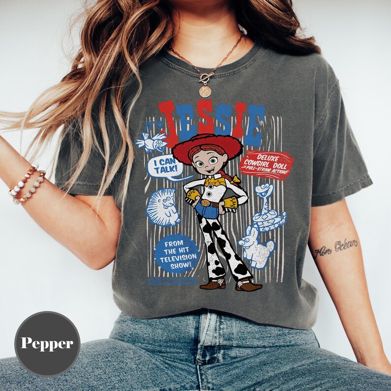 May include: Dark grey t-shirt featuring a cartoon image of Jessie from Toy Story. The graphic includes the text "JESSIE", "I CAN TALK!", and "DELUXE COWGIRL DOLL". Additional text reads "FROM THE HIT TELEVISION SHOW!" The shirt has a vintage, worn look.