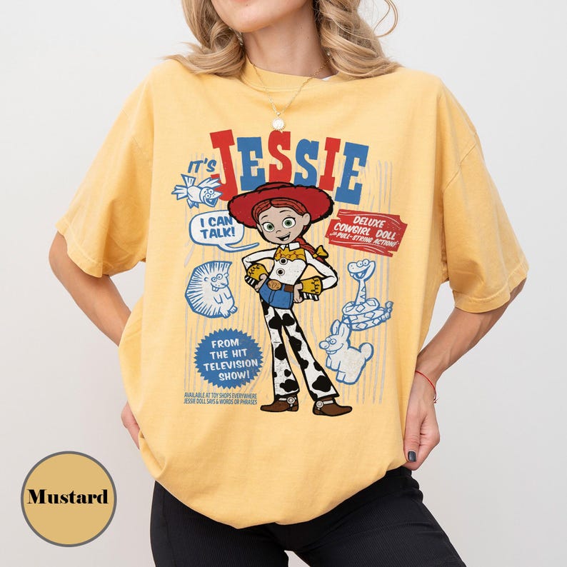 May include: Mustard-colored t-shirt featuring a graphic of the character Jessie from the Toy Story franchise. The design includes the text "It's Jessie," "I Can Talk!", and "Deluxe Cowgirl Doll - Pull-String Action!" with additional cartoon illustrations.