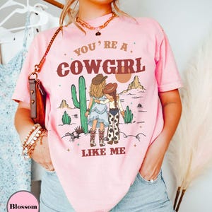 May include: A light pink short-sleeve t-shirt featuring a graphic of two figures in cowboy hats standing in a desert landscape with cacti and mountains. The text reads YOU'RE A COWGIRL LIKE ME in brown lettering. The shirt has a crew neckline.