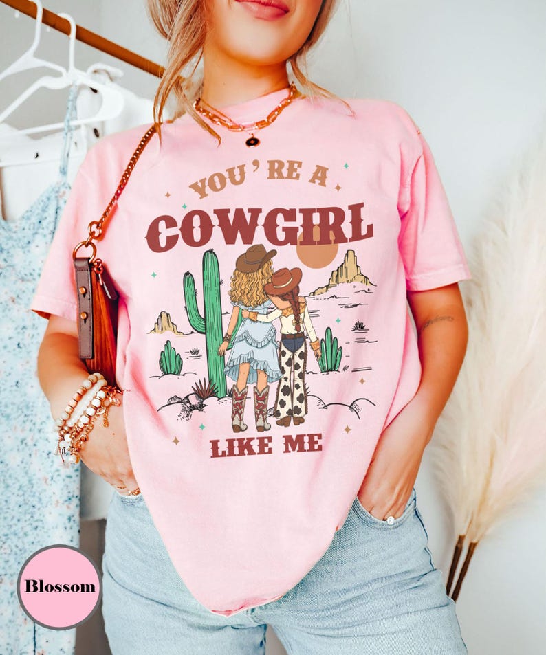 May include: A light pink short-sleeve t-shirt featuring a graphic of two figures in cowboy hats standing in a desert landscape with cacti and mountains. The text reads YOU'RE A COWGIRL LIKE ME in brown lettering. The shirt has a crew neckline.