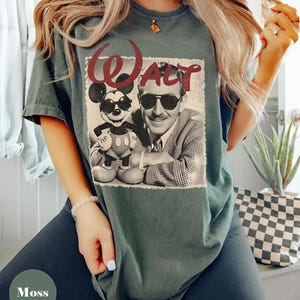 May include: Moss green t-shirt featuring a vintage-style graphic of Walt Disney and Mickey Mouse wearing sunglasses. The word "Walt" is printed above the image in a burgundy script font. The shirt is a relaxed fit.