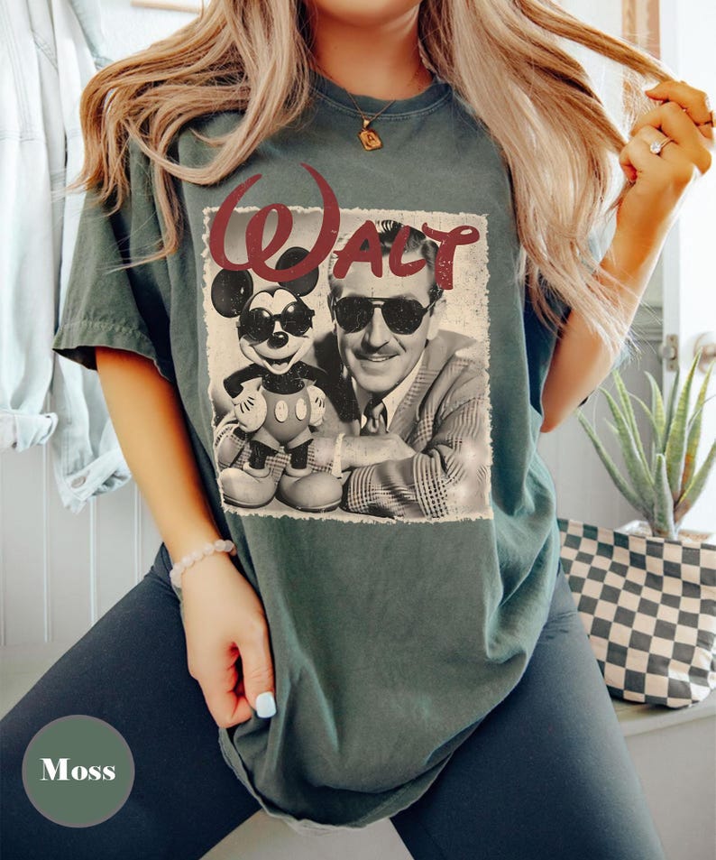 May include: Moss green t-shirt featuring a vintage-style graphic of Walt Disney and Mickey Mouse wearing sunglasses. The word "Walt" is printed above the image in a burgundy script font. The shirt is a relaxed fit.