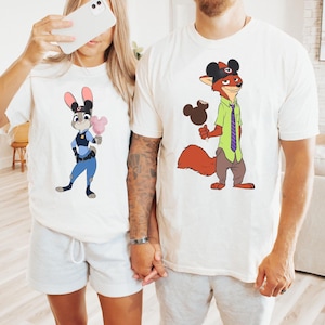 Puede incluir: Dos camisetas blanquecinas con personajes de dibujos animados. Una camiseta muestra un conejo con uniforme de polic&iacute;a sosteniendo algod&oacute;n de az&uacute;car. La otra camiseta muestra un zorro con un helado de Mickey Mouse. Ambas camisetas son de manga corta.