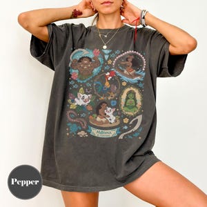 May include: Dark gray t-shirt featuring a colorful graphic of characters from the animated film "Moana." The design includes images of Maui, Moana, Hei Hei, Pua, and other characters. The words "Moana & Friends" are also visible.