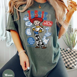 May include: A moss green t-shirt featuring a graphic of Jessie from Toy Story. The design includes the word "Jessie" in red and blue, along with cartoon images and the text "I CAN TALK!" and "DELUXE COWGIRL DOLL".