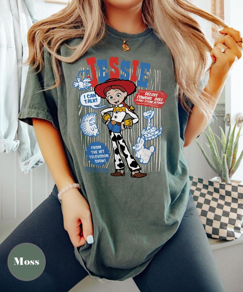 May include: A moss green t-shirt featuring a graphic of Jessie from Toy Story. The design includes the word "Jessie" in red and blue, along with cartoon images and the text "I CAN TALK!" and "DELUXE COWGIRL DOLL".