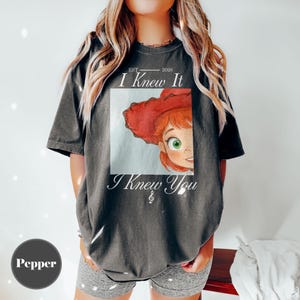 May include: A charcoal gray short-sleeve t-shirt featuring a graphic print of a cartoon character with red hair and a red hat. The text reads EST 2026 I Knew It I Knew You with a small musical note symbol below the text. The shirt has a loose fit.